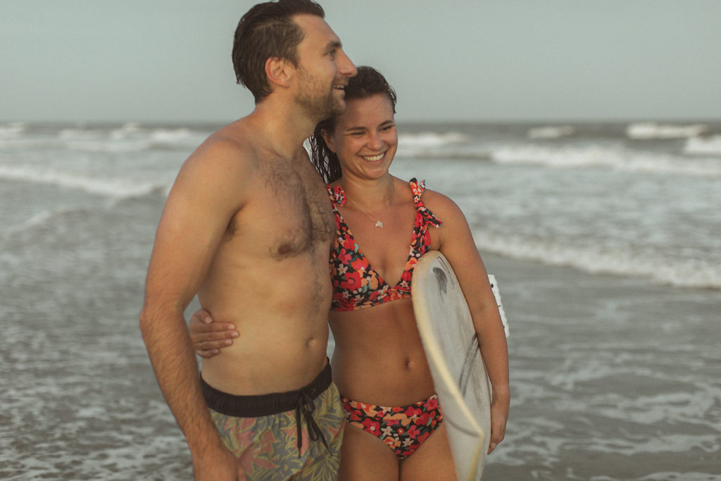 man and woman hugging each other while on the beach and holding a surfboard for their outdoor engagement photos with Virginia engagement photographer