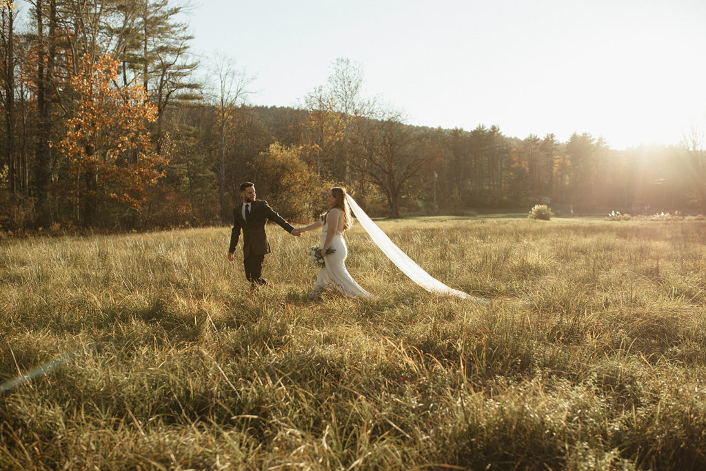 Charlottesville wedding photographer captures bride and groom holding hands and walking through a field together at sunset as the groom leads the bride
