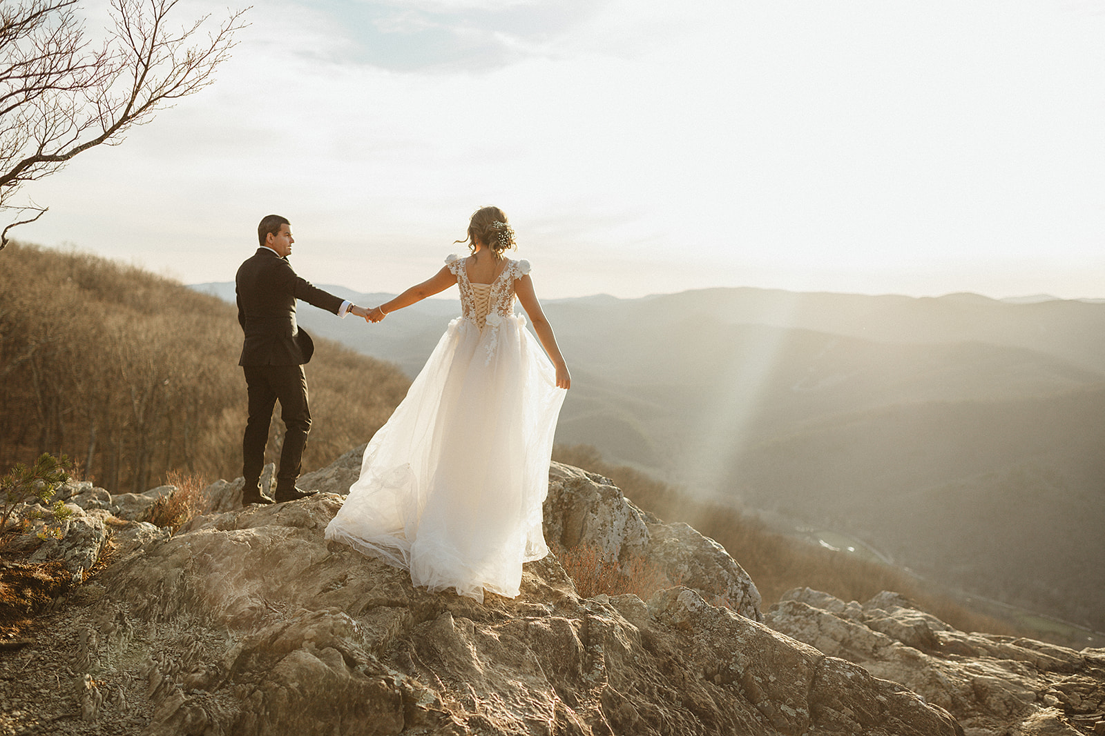bride and groom holding hands and hiking up a mountains together as the sun shines down at them captured by Virginia wedding photographer