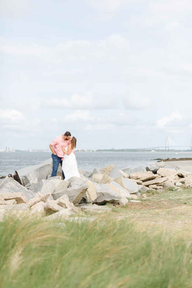 An engaged couple shares a tender kiss on the rocky shores of Sullivan's Island, capturing a moment of love against a backdrop of expansive skies and distant bridge views.