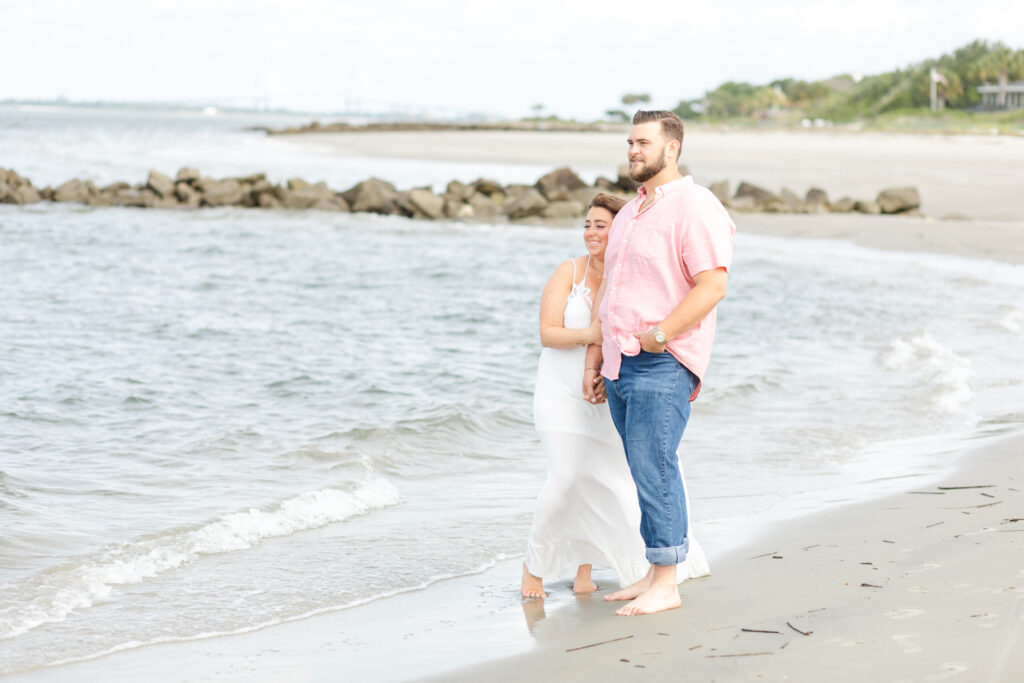 A couple enjoys a peaceful moment during their Sullivan's Island engagement, standing barefoot on the sandy beach with gentle waves and a rocky jetty in the background.