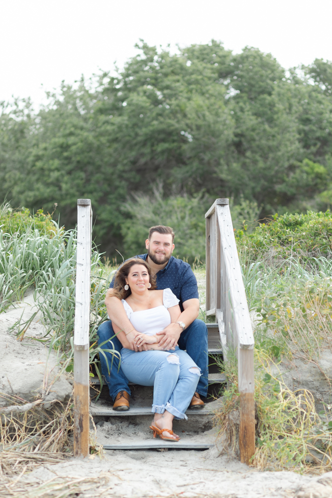 A joyful couple poses for their Sullivan's Island engagement photo, sitting together on wooden steps amidst beach greenery, exuding casual elegance and happiness.