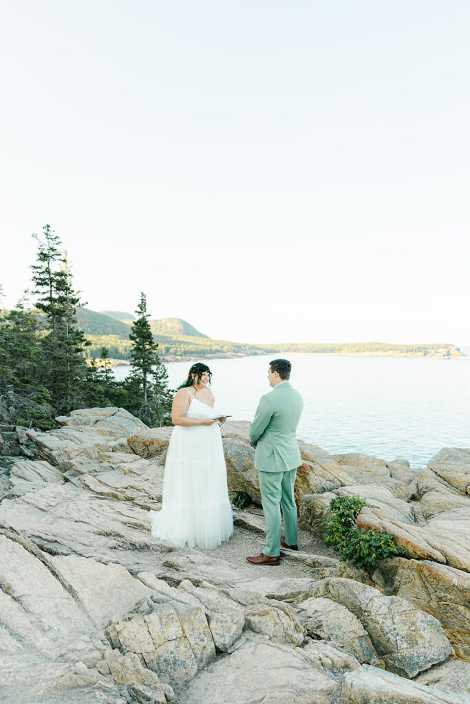 A couple shares their vows on the rugged, rocky landscape of Otter Cliffs in Acadia National Park, with sweeping views of the ocean and distant forested mountains in the background.