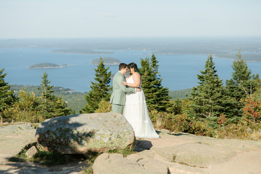 A couple embraces on Cadillac Mountain, one of the many stunning locations in Acadia National Park, surrounded by evergreen trees and sparkling blue waters in the distance.