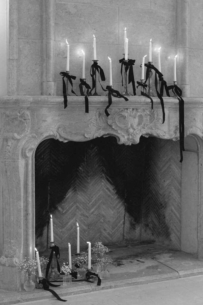 Black and white image of an ornate fireplace adorned with tall candlesticks wrapped in black velvet ribbons. Additional candles and small floral arrangements decorate the hearth, creating a moody, romantic atmosphere.