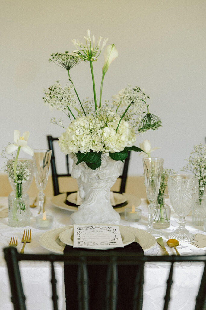 Elegant, non-traditional wedding table centerpiece featuring a classical statue vase filled with white hydrangeas, delicate greenery, and minimalist floral accents. Vintage glassware and gold flatware complement the modern yet timeless design.