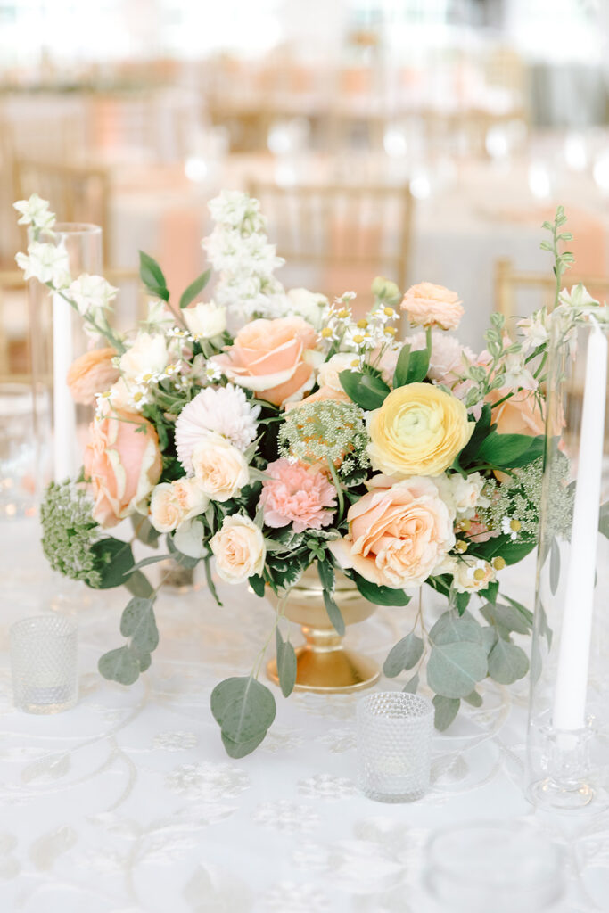 Vibrant wedding table centerpiece featuring soft peach and yellow roses, white ranunculus, and lush greenery. The arrangement is displayed in a gold vase, complemented by candle votives on a lace tablecloth for an airy, romantic look.