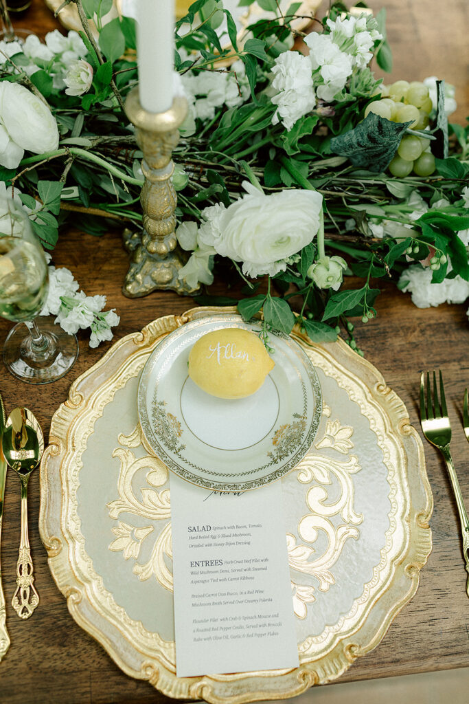 Elegant table setting with a gold and white vintage plate, delicate floral accents, and a personalized lemon place card. Lush greenery and white flowers surround gold candlesticks, creating a fresh, sophisticated design.