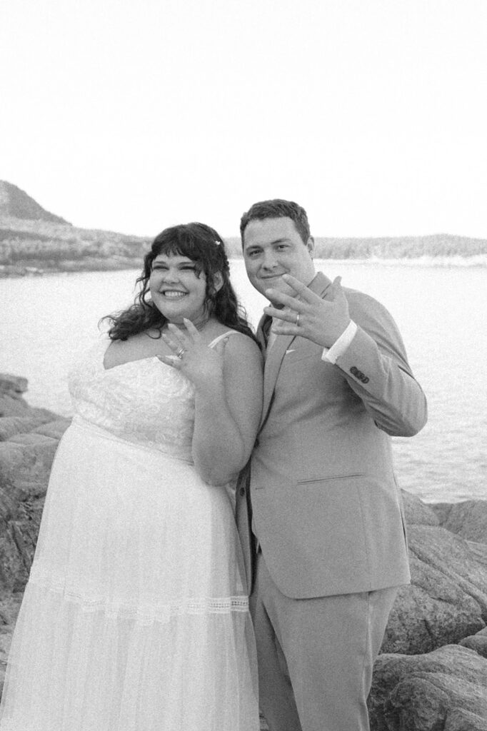 A newlywed couple smiling and showing off their wedding rings while standing on rocky cliffs with the ocean in the background