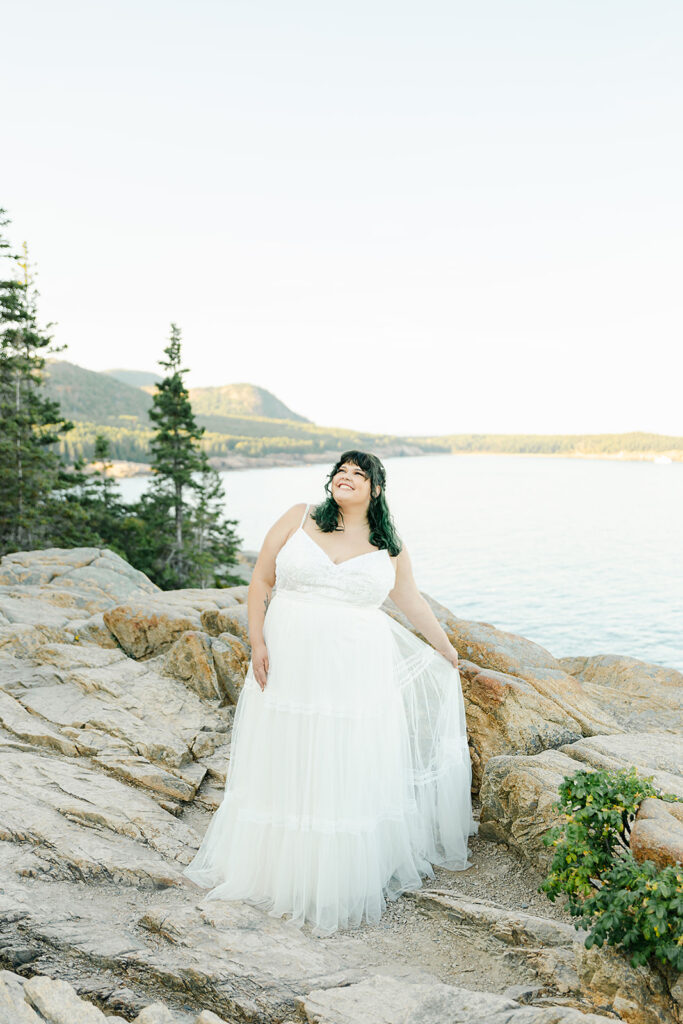 A bride stands on the rocky cliffs of Acadia National Park, looking out at the serene ocean and distant mountains during her elopement