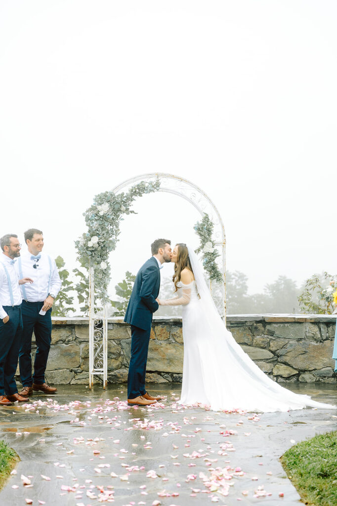 Newlyweds sharing their first kiss under a floral arch on a misty day at Bluemont Vineyard, surrounded by their wedding party.