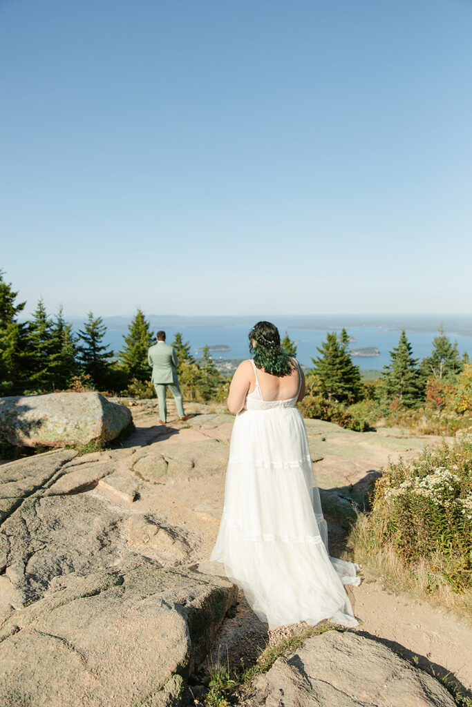 The bride walks towards her groom for their first look, surrounded by rocky terrain and sweeping ocean views on Cadillac Mountain in Acadia National Park