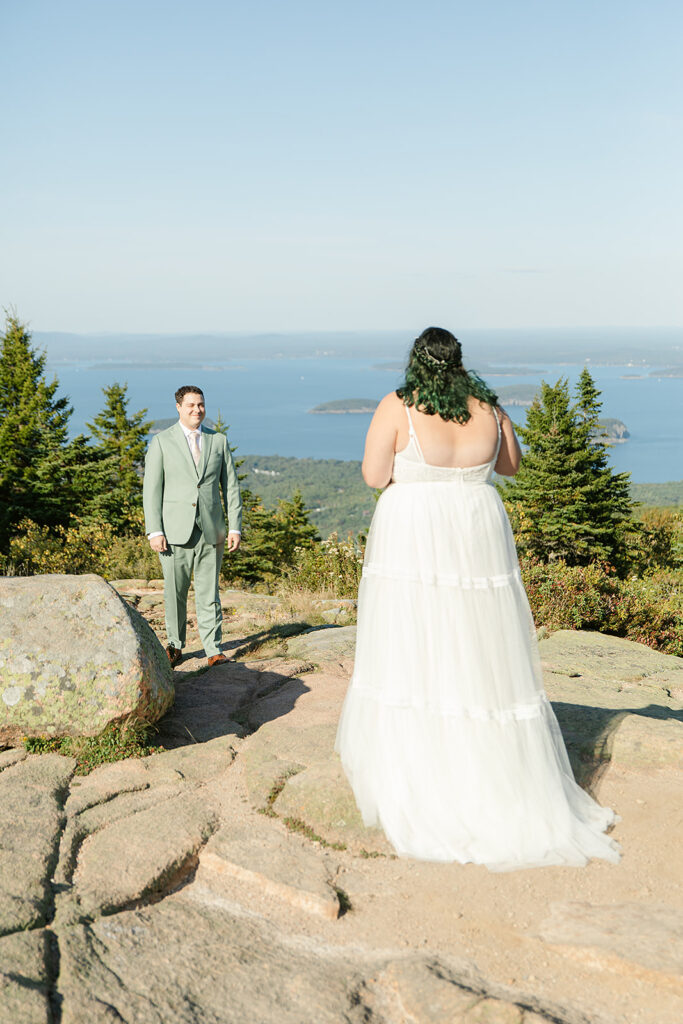 Estella and Calvin’s intimate elopement on Cadillac Mountain in Acadia National Park, with the groom smiling as the bride approaches against a breathtaking coastal backdrop