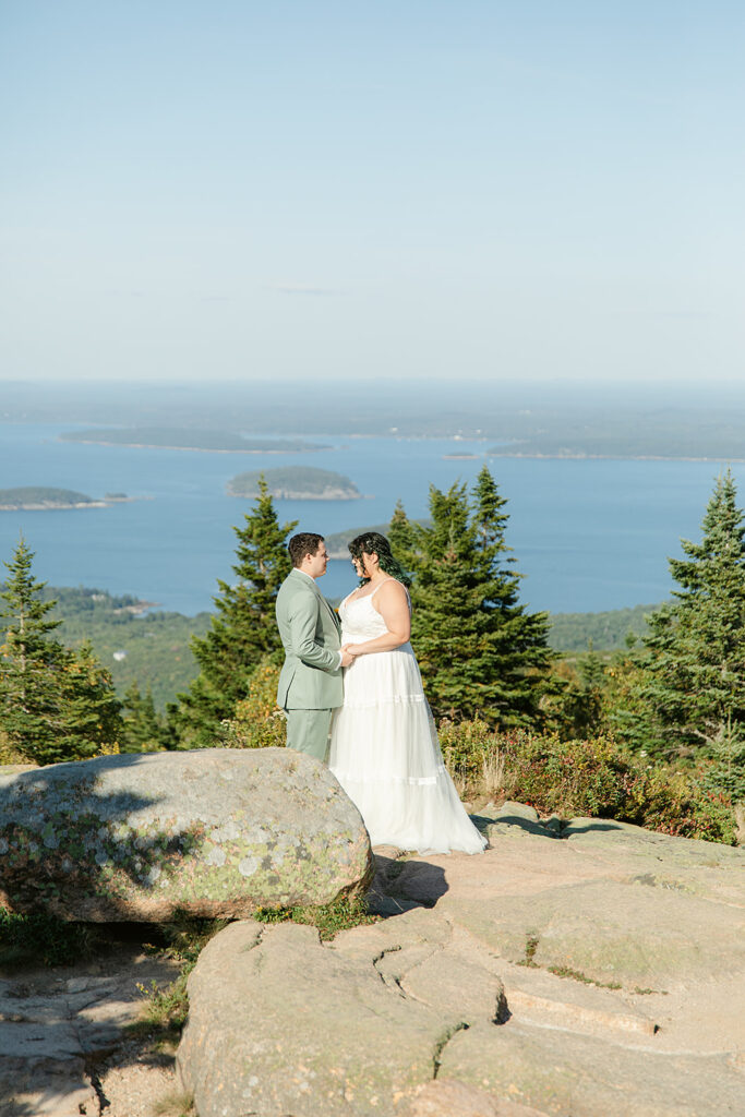 A newlywed couple stands together on Cadillac Mountain in Acadia National Park, surrounded by panoramic views of lush trees and the Atlantic Ocean