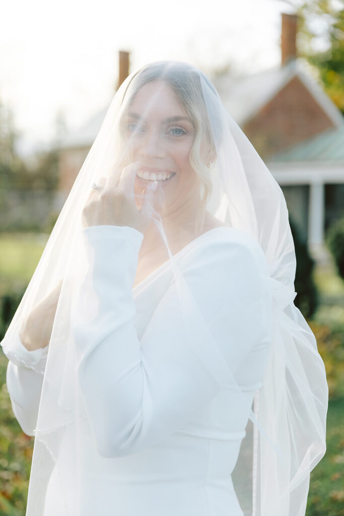 A smiling bride playfully holding her veil over her face while standing in the sunlight at Chatham Manor in Fredericksburg, Virginia.