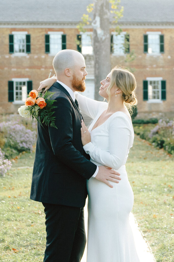  A newlywed couple embracing in front of Chatham Manor in Fredericksburg, Virginia, with the groom holding a bouquet of orange roses and the bride looking up at him lovingly.