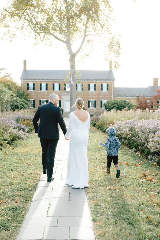 A couple in wedding attire walking hand-in-hand with their young child along a garden path at Chatham Manor in 