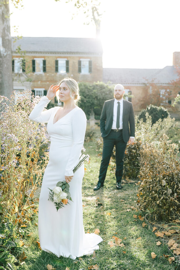 A bride in a long-sleeved white gown holding a bouquet as she gazes into the distance, with her groom standing in the background at Chatham Manor in Fredericksburg, Virginia.