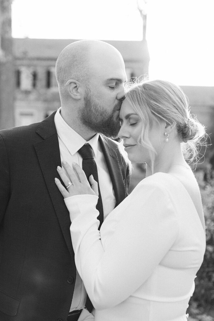 A black-and-white photo of a groom kissing his bride’s forehead while she smiles softly, standing together at Chatham Manor in Fredericksburg, Virginia.