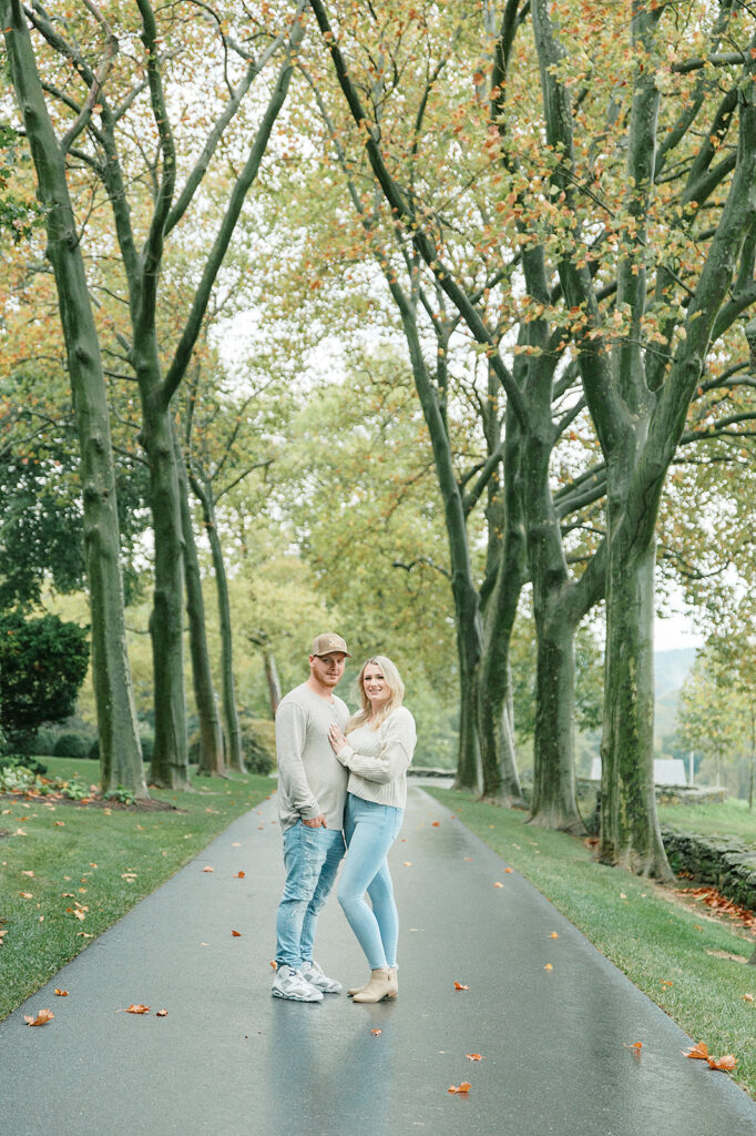 Romantic portrait of a couple standing on a pathway surrounded by tall trees with autumn leaves.