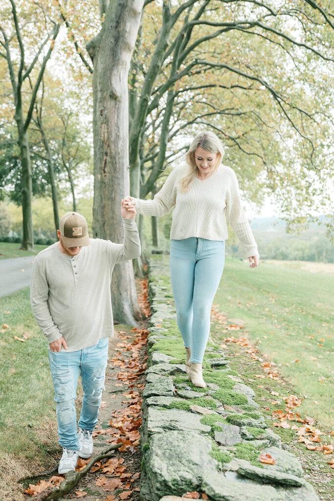 Couple holding hands as they walk along a moss-covered stone wall surrounded by lush trees at Drumore Estate.