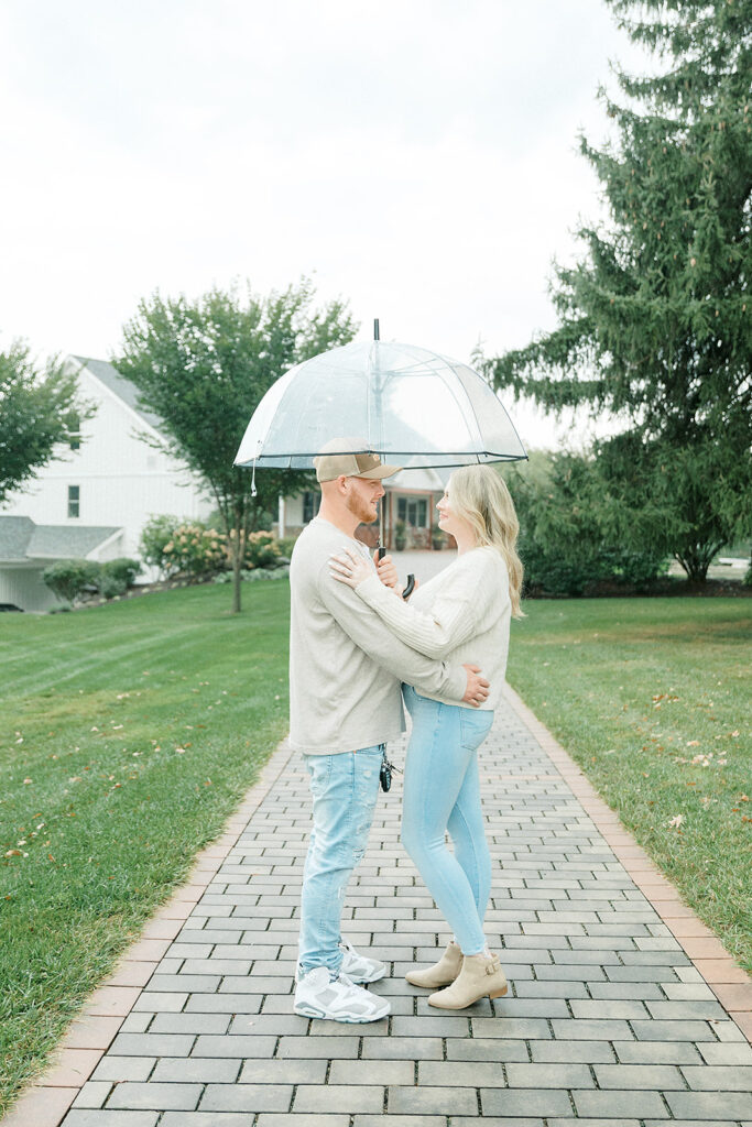 A couple holding each other under a clear umbrella on a rainy day at Drumore Estate’s brick pathway.