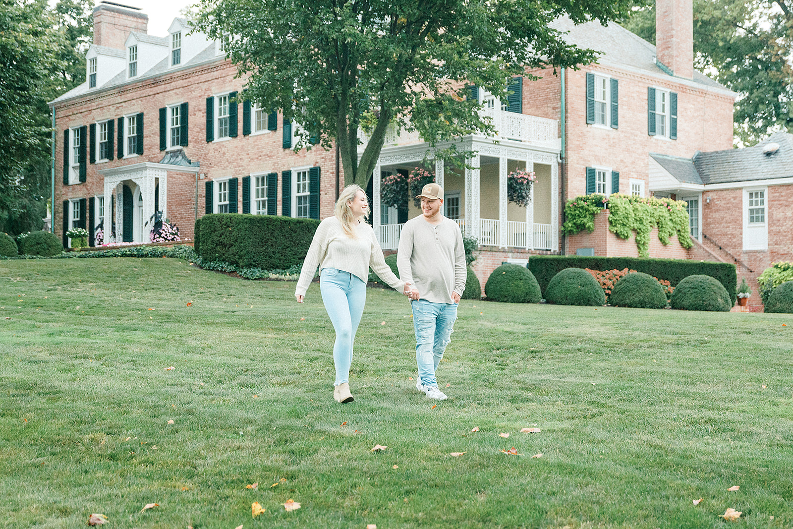 A couple holding hands and walking across the green lawn in front of the historic Drumore Estate in Lancaster, PA, during their engagement session.