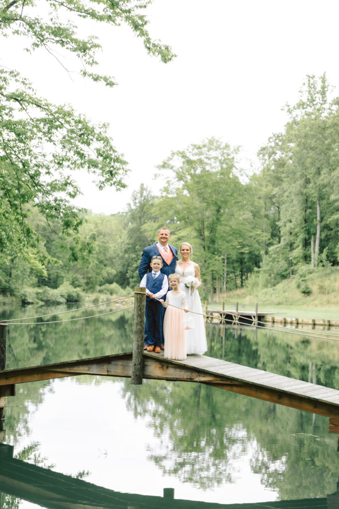 Bride, groom, and their children standing on a dock by a peaceful pond at Layz S Ranch, surrounded by lush greenery.