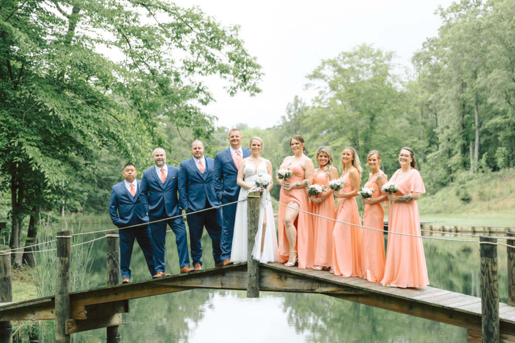 Bride, groom, bridesmaids in coral dresses, and groomsmen in navy suits standing on a wooden bridge at Layz S Ranch, with a serene pond and lush greenery in the background, showcasing the venue’s rustic charm and natural beauty.