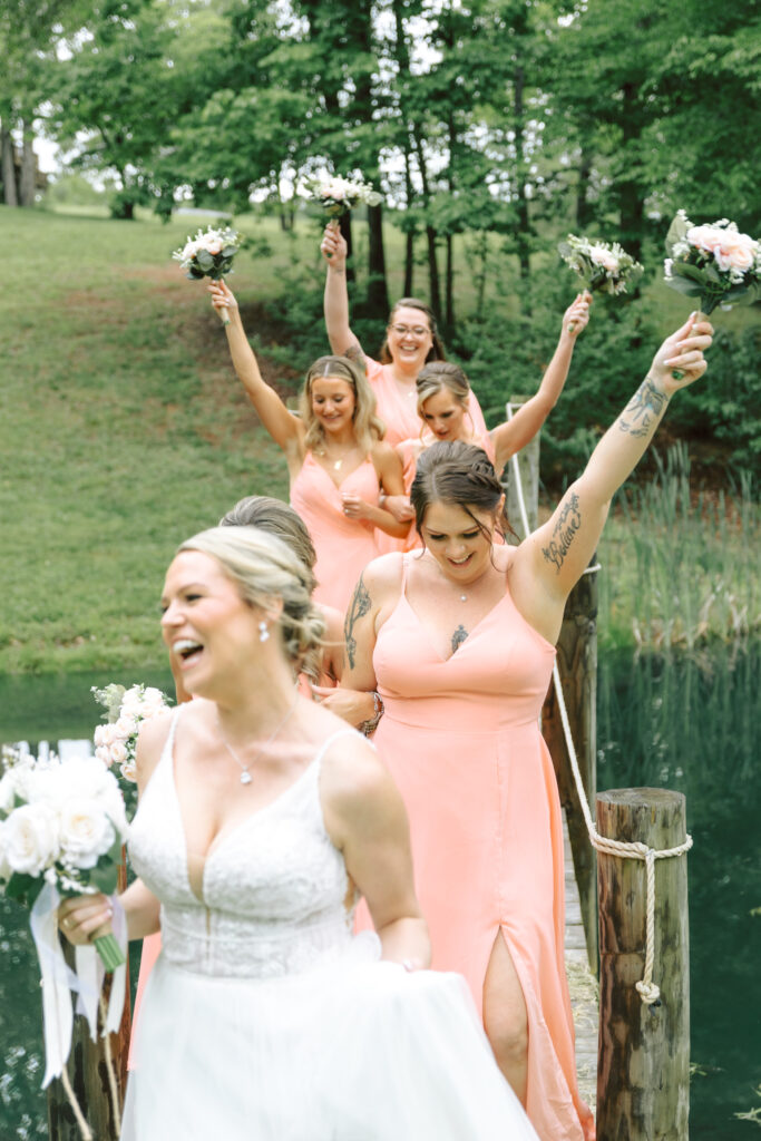 Bride laughing with her bridesmaids in coral dresses as they cross a wooden bridge at Layz S Ranch, holding bouquets in celebration.