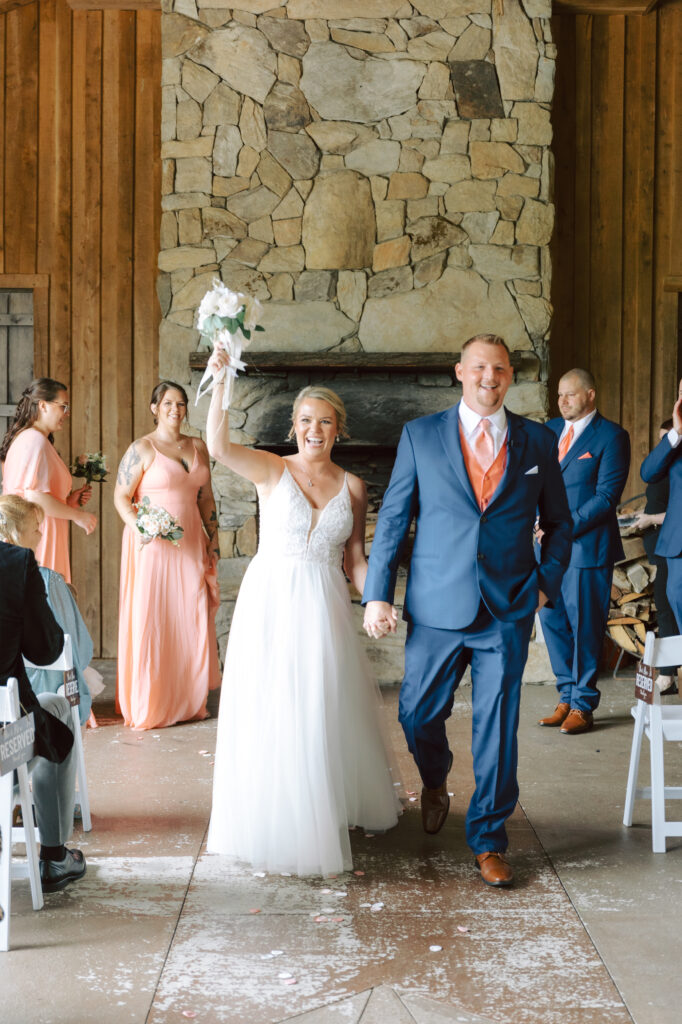 Bride and groom exiting their ceremony at Layz S Ranch, with the bride raising her bouquet in celebration in front of a stone fireplace.