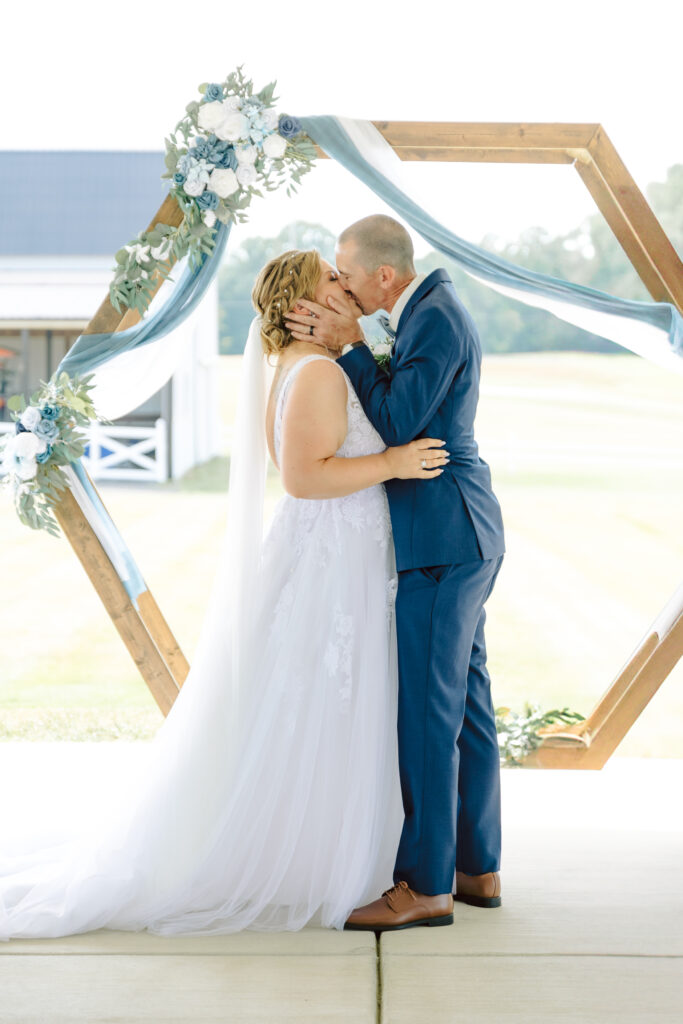 Bride and groom share their first kiss during their Bowles Farm wedding ceremony under a hexagonal arbor adorned with blue and white floral decor. The rustic setting highlights the charm of this Southern Maryland venue, perfect for outdoor celebrations.