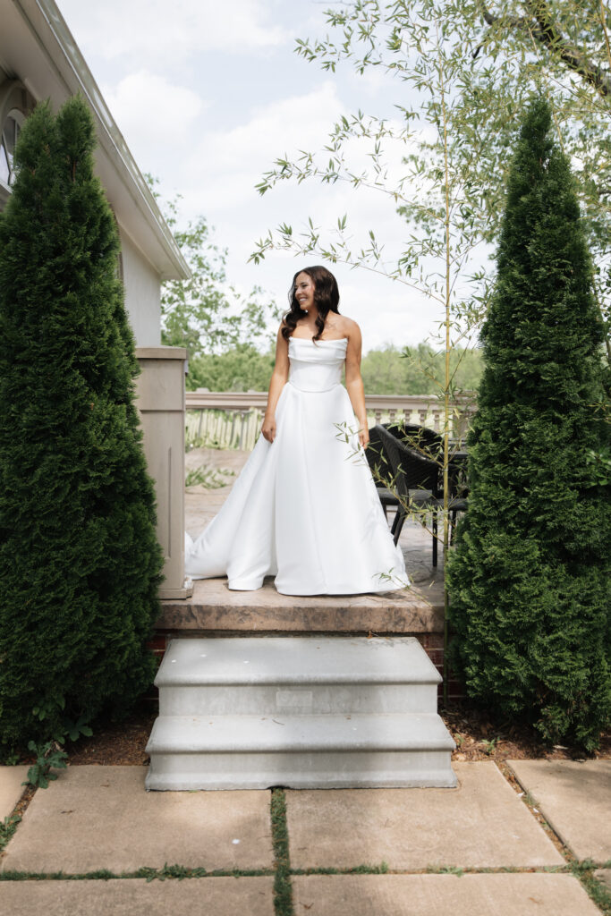 Bride smiling in a classic off-the-shoulder white gown standing between lush green topiary trees at Morais Vineyards.