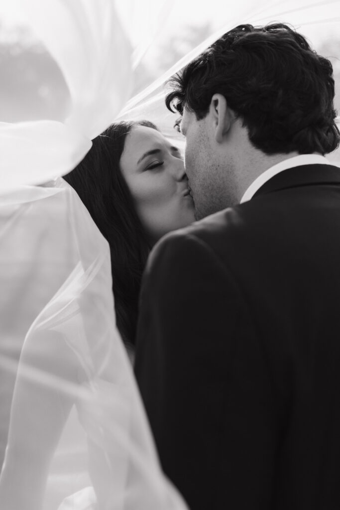 Intimate black-and-white kiss under the veil, bride’s hand gently holding her groom's lapel.

