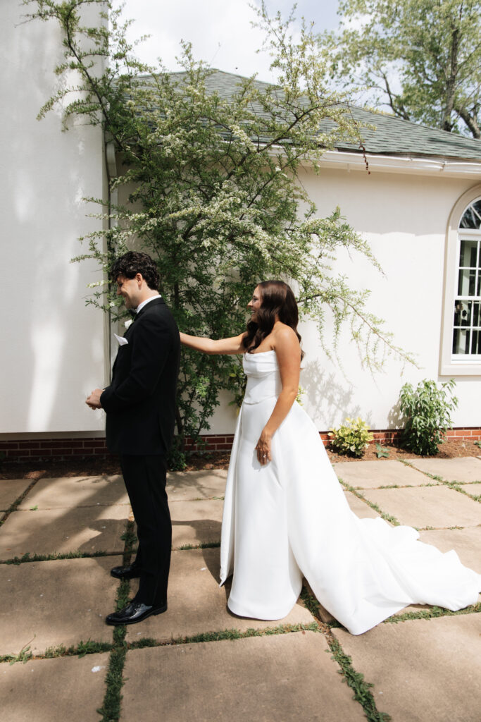 Bride tapping groom’s shoulder during emotional first look beside the chapel wall.
