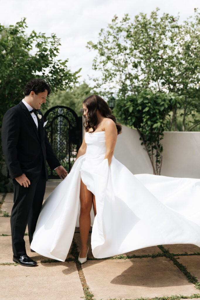 Groom admiring bride’s dress as she lifts it playfully to show a bold slit in the skirt.

