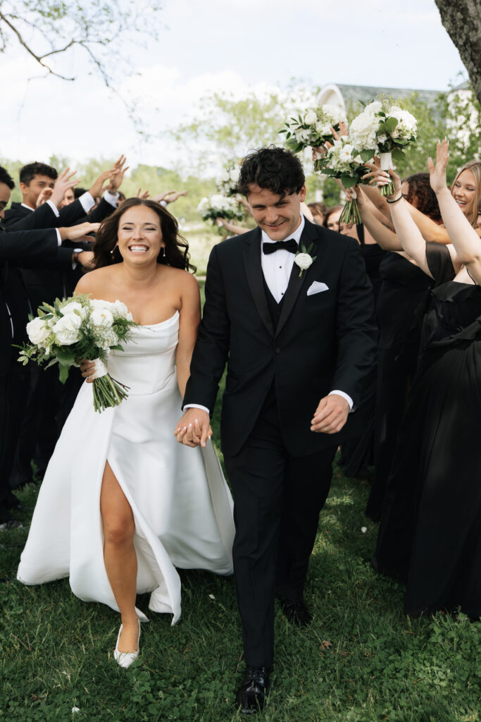 Bride and groom laughing while walking hand in hand as their black-tie wedding party cheers around them at Morais Vineyards in Virginia